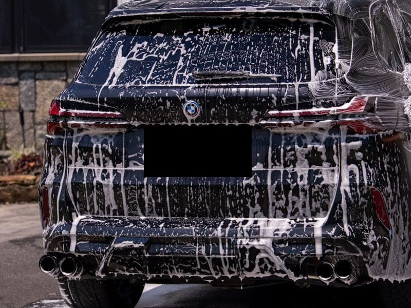 Rear of a matte-black BMW X5 covered in wash foam in a residential driveway, owner-operator's arm with wash mitt at the right of frame, license plate obscured.