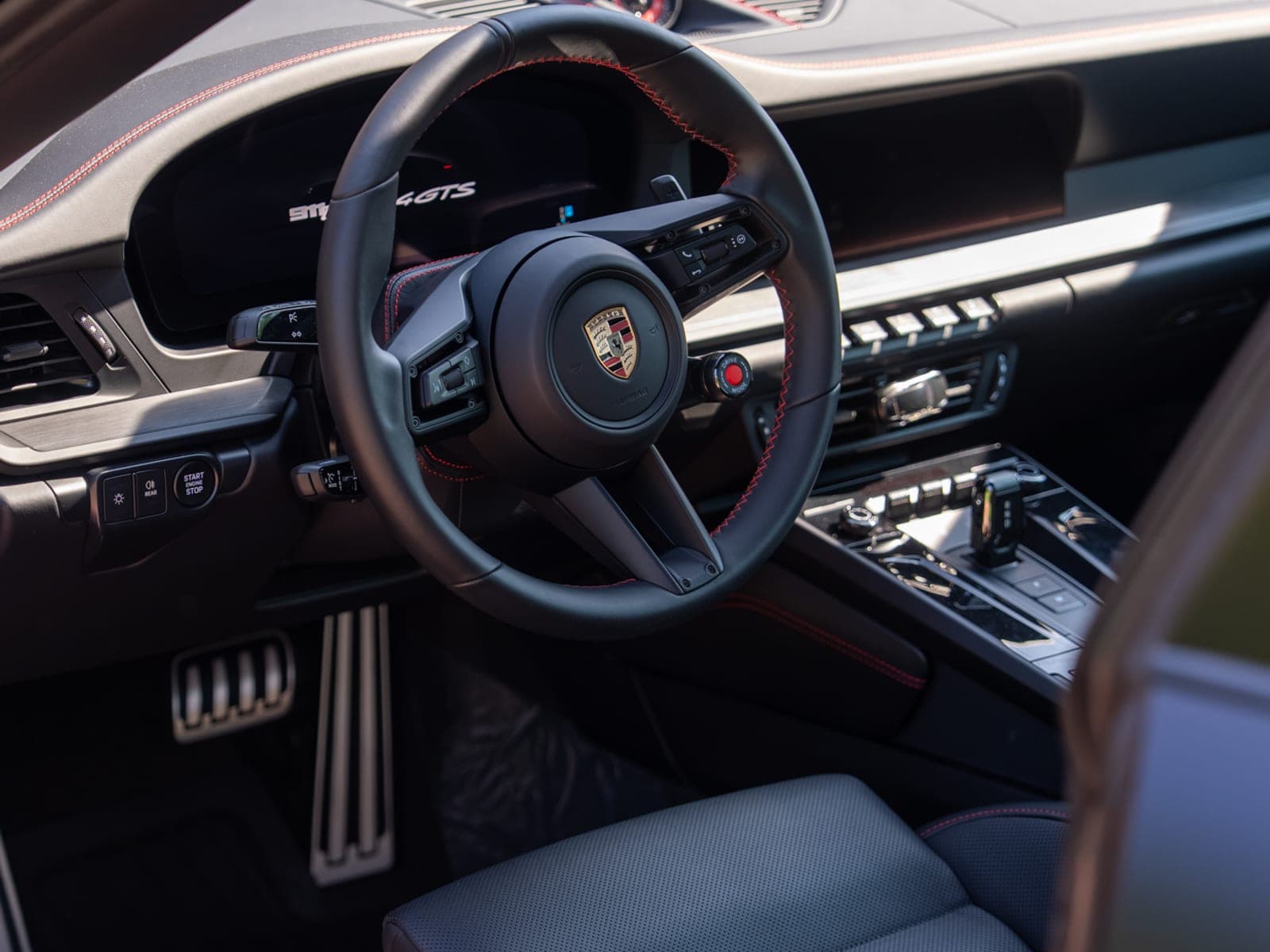 Interior of a Porsche 911 Targa 4 GTS: red-stitched steering wheel, dashboard, aluminum pedals, and cloth seat in natural daylight.