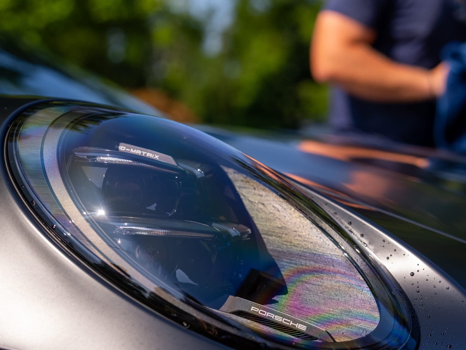 Water beaded across a matte-black Porsche 911 headlight and adjacent panel after a wash, iridescent reflection in the LED housing, Pro Performance owner-operator softly out of focus behind the hood.